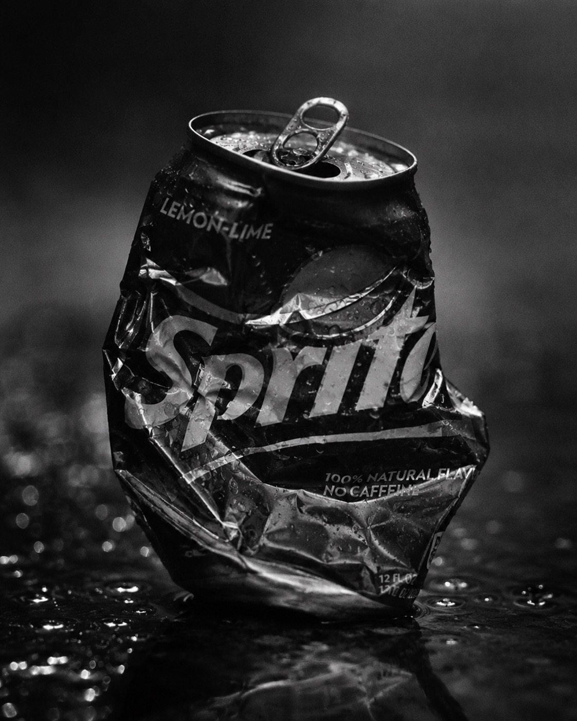 A black and white photograph of a crushed Sprite can on a wet surface.